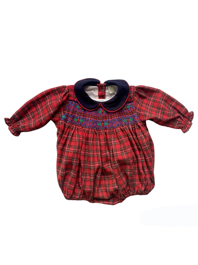 A red tartan romper with a navy contrast collar and hand smocking featuring holly embroidery