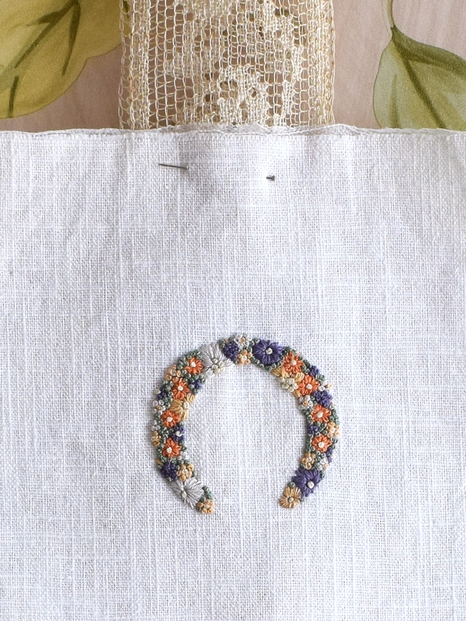 An embroidered Sunshine Garden Horseshoe, of Golden Yellows and Bright Orange Blossoms with Green French Knot grass background.  Pinned to a wide lace ribbon.