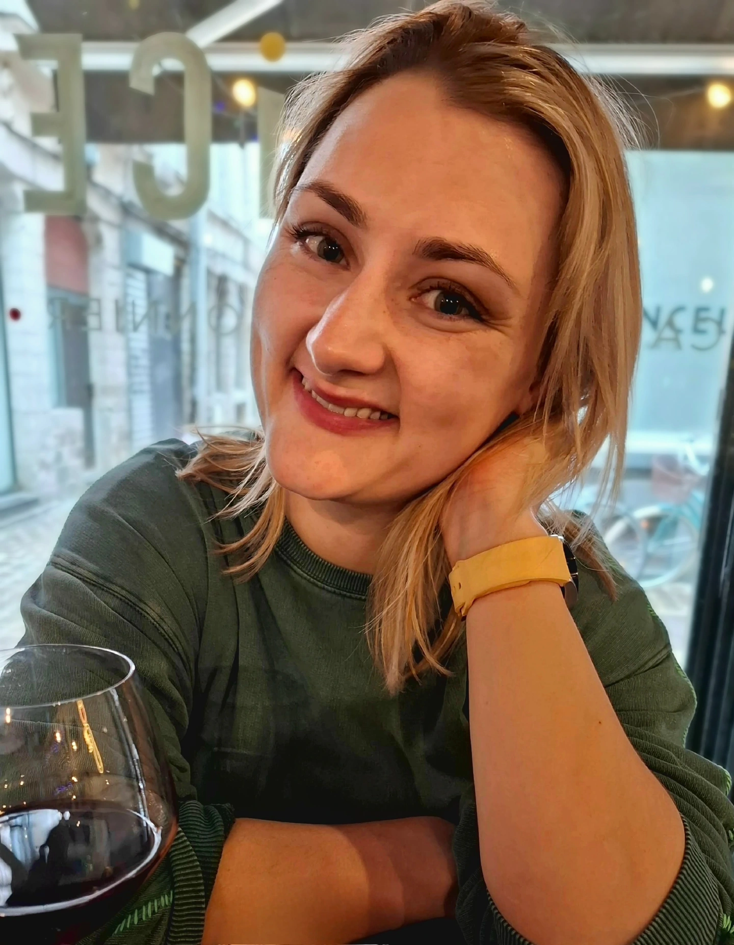 Up close photo of a caucasian woman with dark blonde hair wearing a green jumper. She is smiling looking at the camera, and there is a large window behind her.  