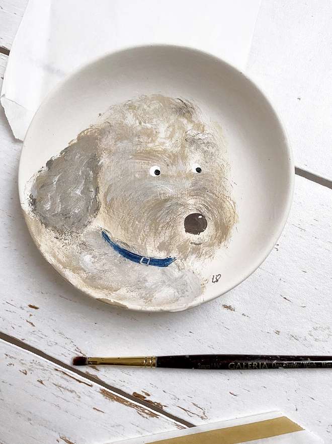 little plate painted with a dog next to a paintbrush and paper bag