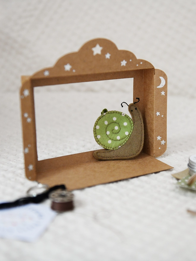 A cardboard mini puppet theatre stands on a white fabric surface with a felt snail inside.