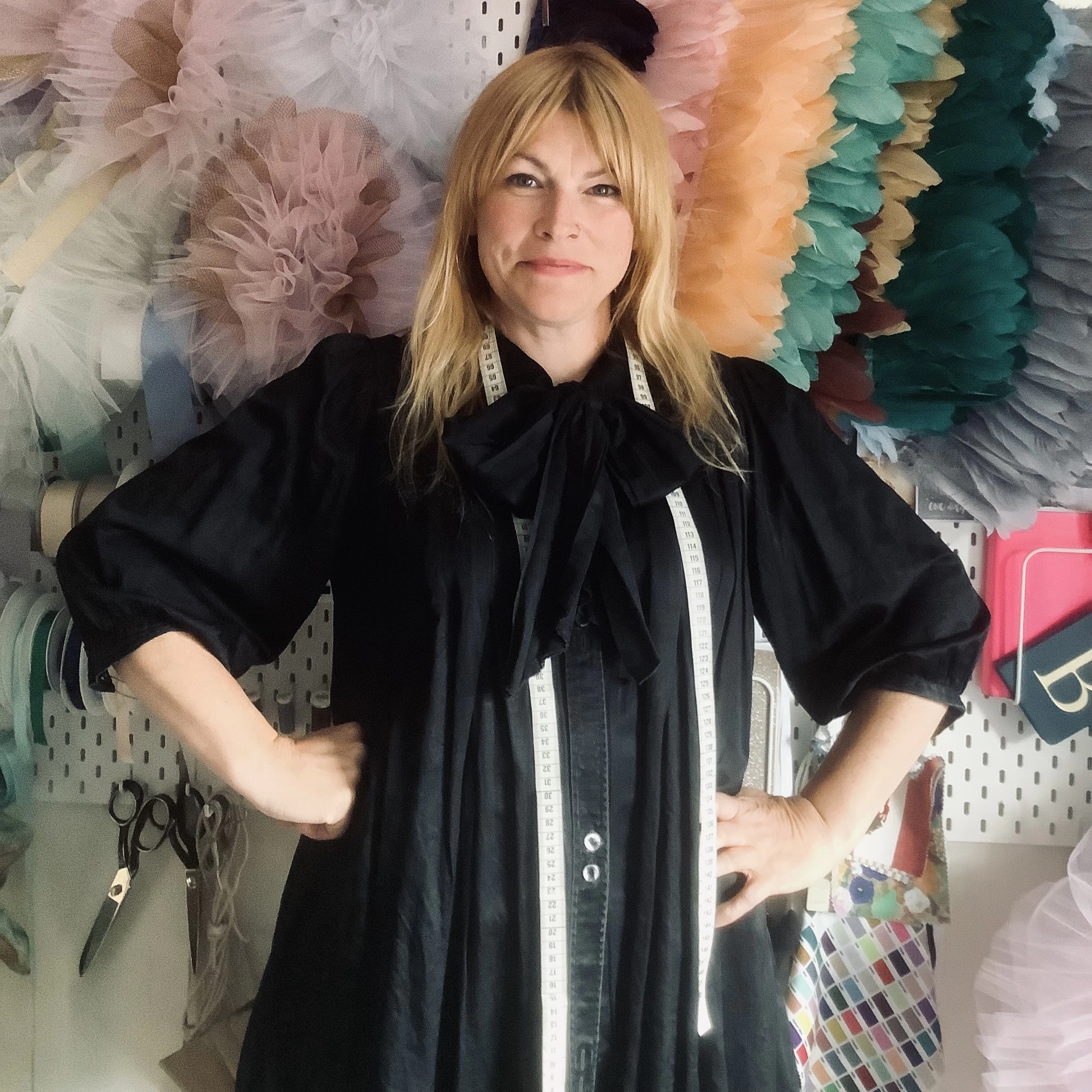 Woman with strawberry blonde hair wearing a black dress with a large bow, and a tape measure draped around the neck.  Standing in her studio surrounded by colourful feathers and tulle