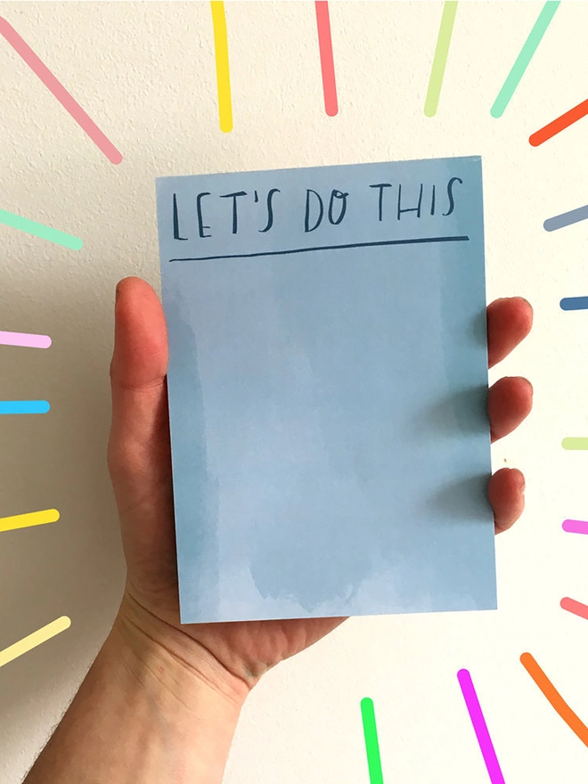 Hand holding desk jotter which reads 'LET'S DO THIS' in blue on blue background
