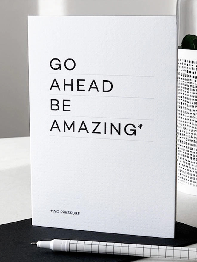 Stood up white card that reads "Go ahead be amazing*" in big letters and "*no pressure" in smaller letters at the bottom in a white themed room.