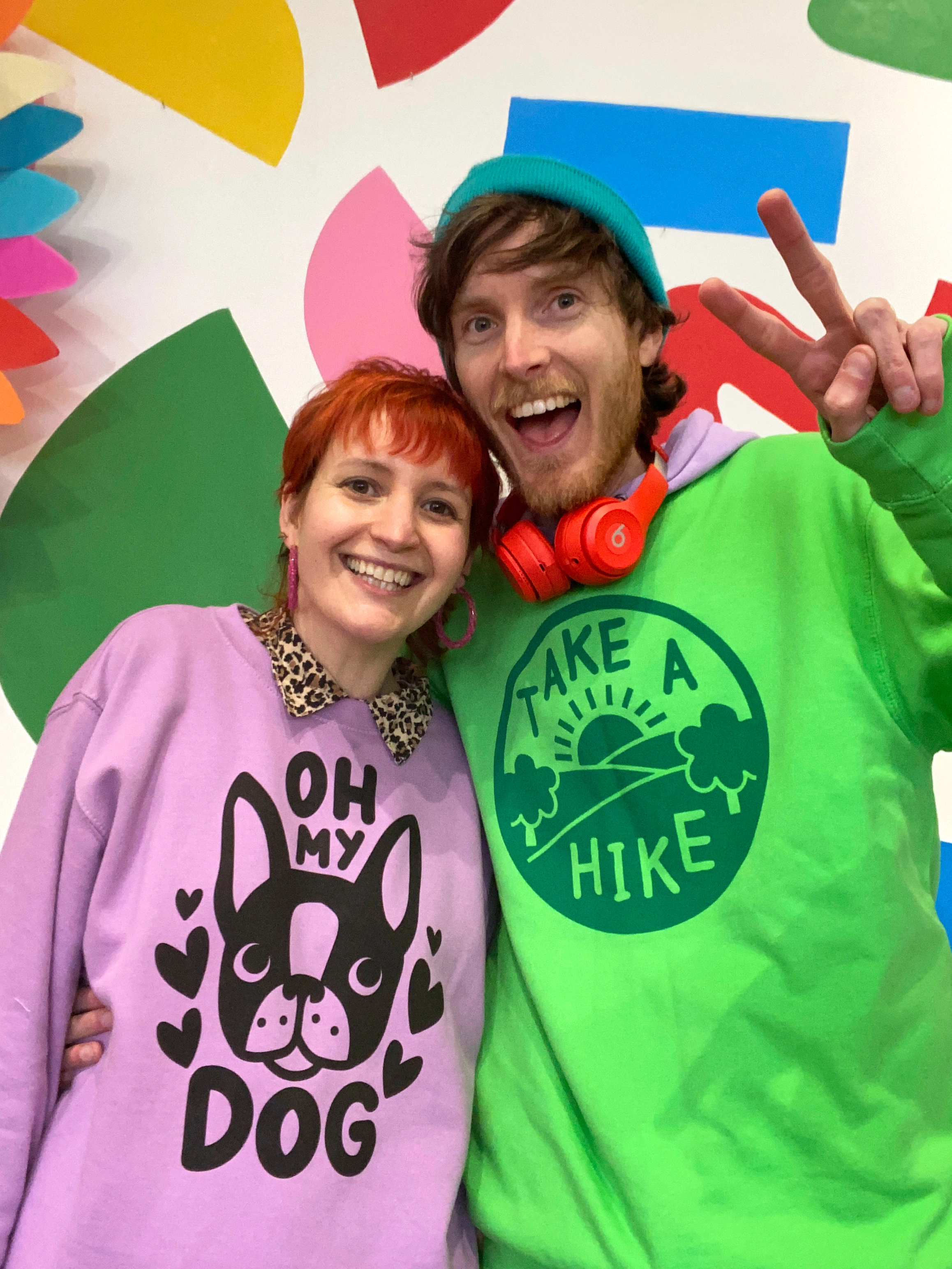 Ali and Jam, the founders of hello DODO Kids, a white woman and white man in their 30's stand against a colourful wall smiling at the camera. They both wear colourful screen printed sweatshirts.