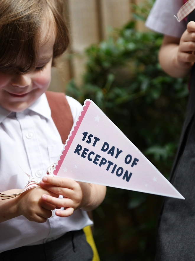 A child in a school uniform holds a pink pennant flag that reads "1st Day of Reception," adorned with ribbons.