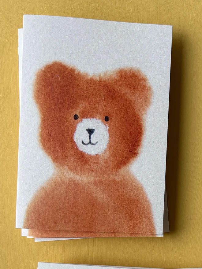 A greetings card featuring a hand painted brown bear cub. The cub is painted in watercolour with a white snout and a friendly smile.