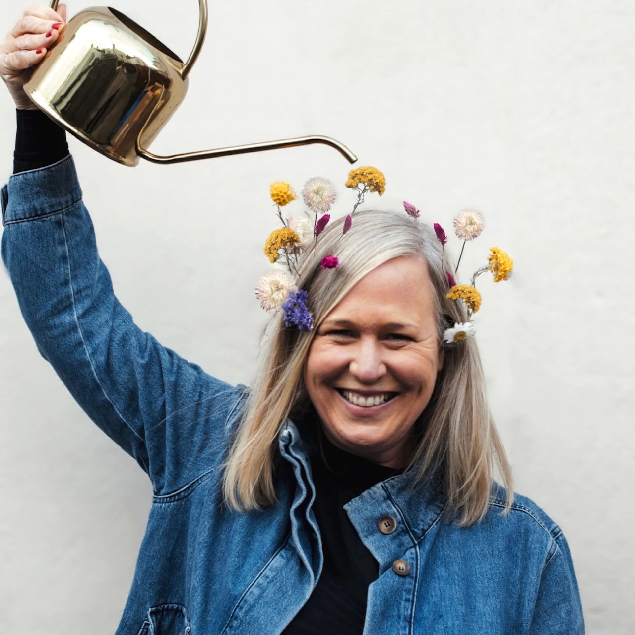 Kate Peers with flowers in her hair holding a metal watering can over her head 