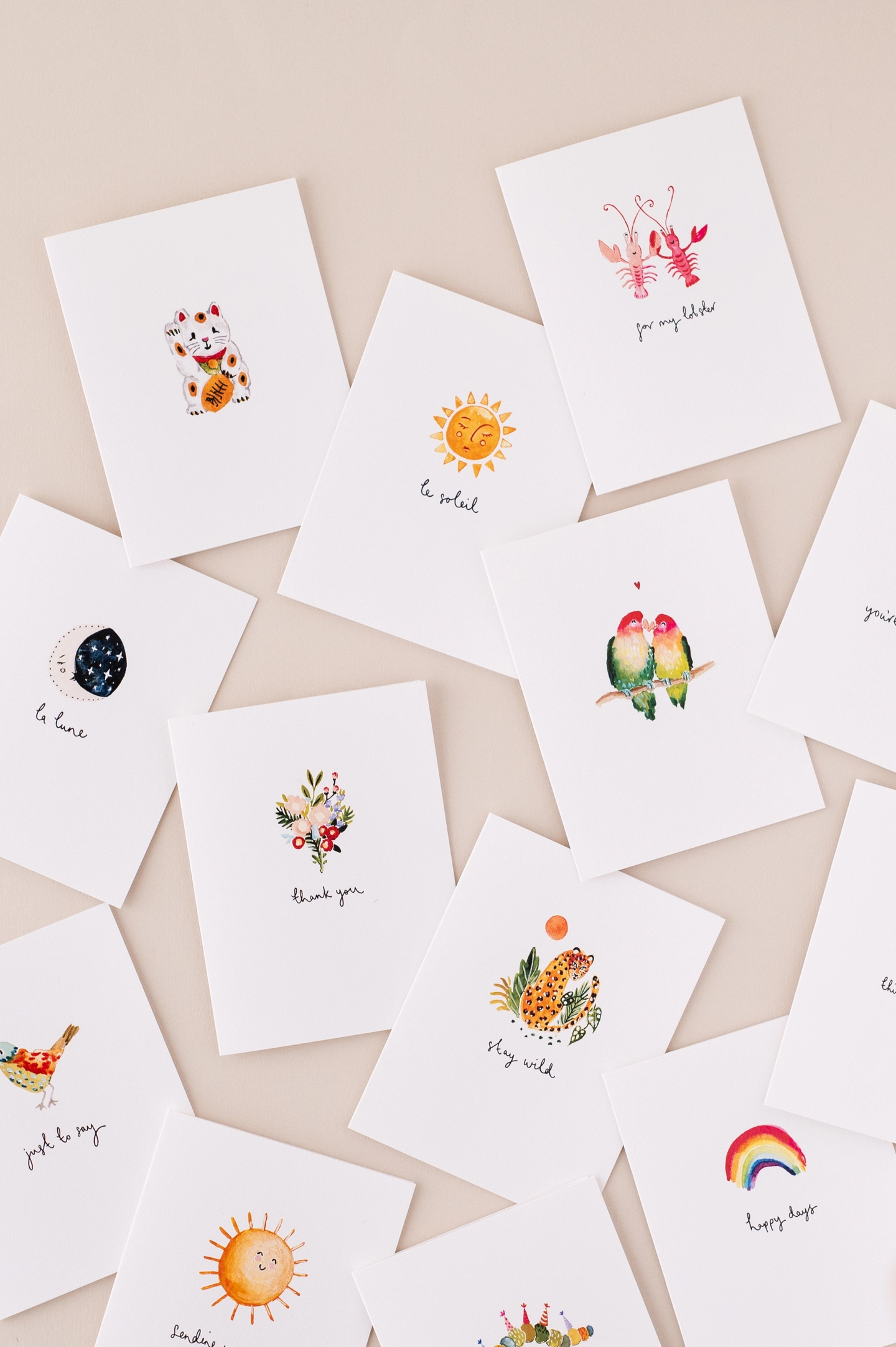 A flat lay photos of Louise Mulgrew's mini size greetings cards, scattered across a neutral pink background. The greetings cards feature colourful gouache motifs with a white background. Illustrations include love birds, a lobster couple, a lucky cat, a bunch of flowers, a rainbow and smiling suns.