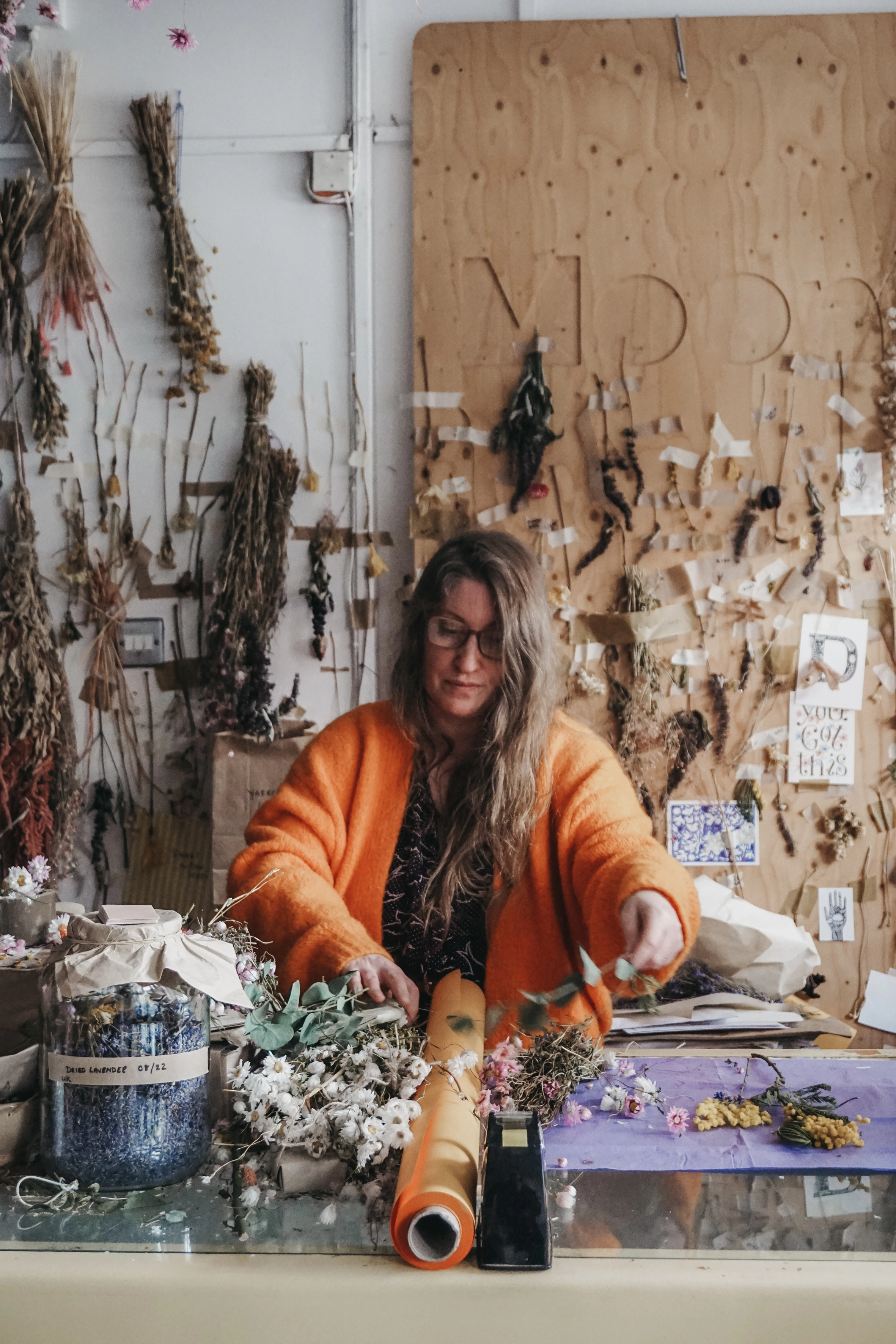 Deborah-Maire Moon standing at a counter, wearing an orange cardigan, making dried flower bouquets.