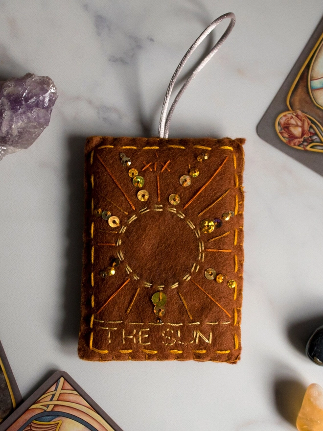 A handcrafted felt ornament with sequins and embroidery depicting "The Sun" tarot card