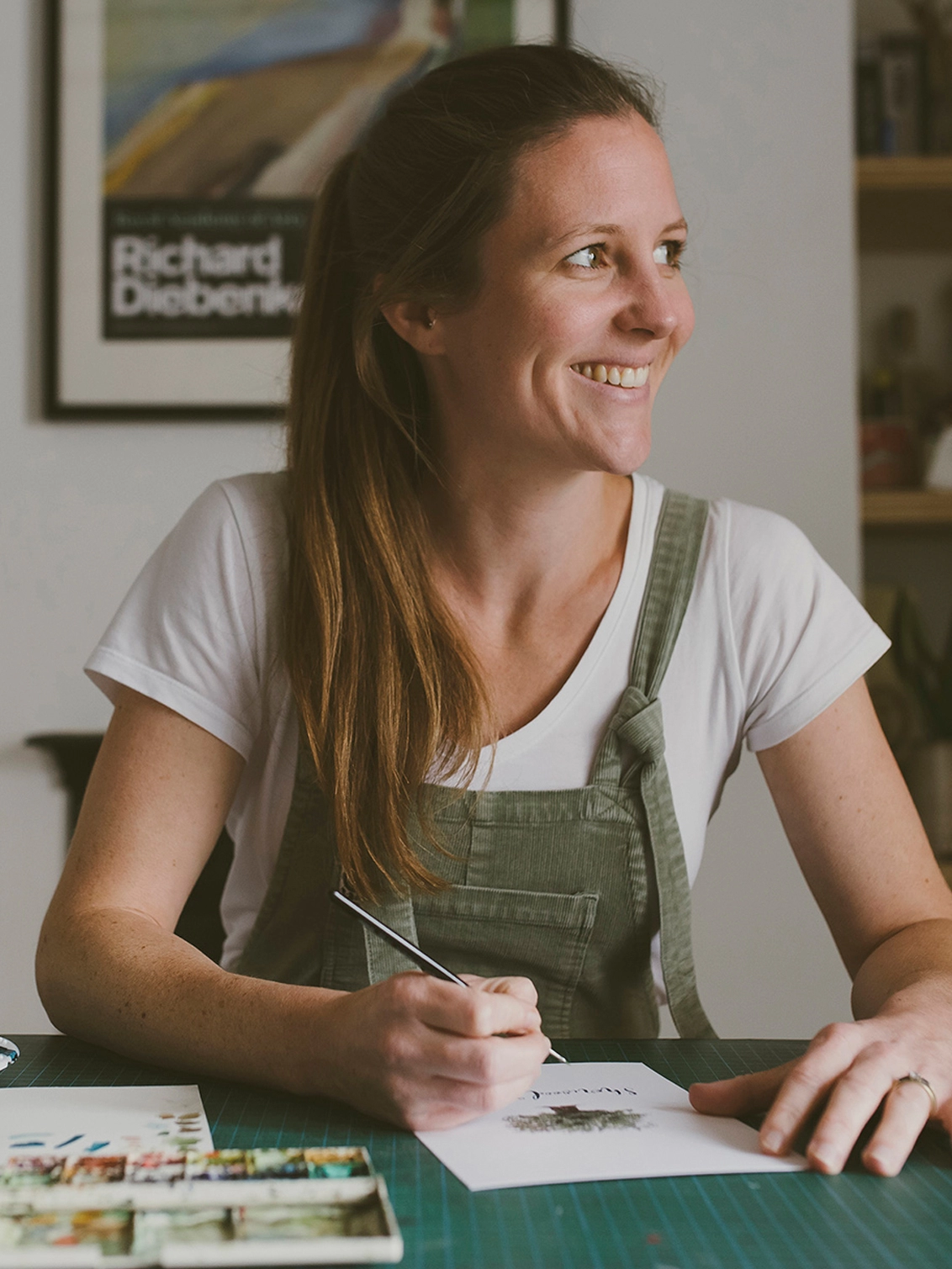 Photo of Imogen Partridge, a white thirty-something female with mid brown hair tied in a pony tail, sitting at a table with her paint brush in her hand. She is looking to the right and smiling, whilst working on a watercolour illustration. She wears olive green dungarees and in the background which is slightly blurred is a white wall with a framed artwork poster on. 