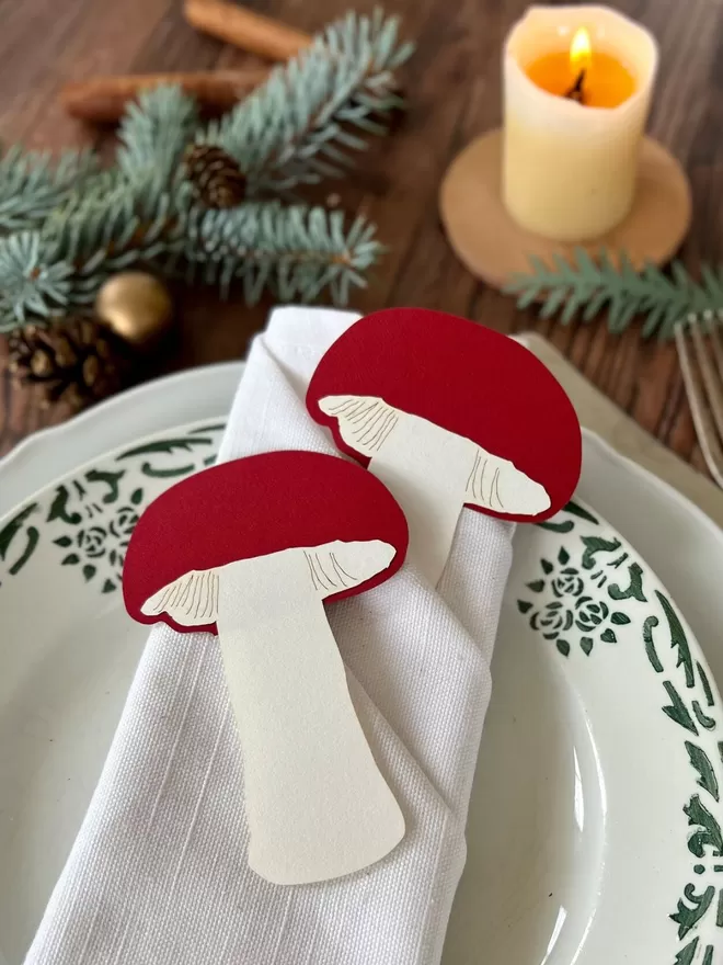 2 red and white paper toadstool decorations resting on top of a napkin and Christmas tableware