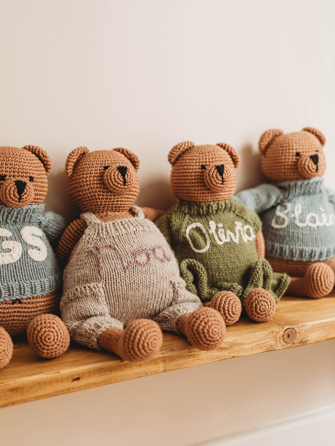 four hand crochet brown teddy bears on wooden shelf wearing hand knit personalised clothing