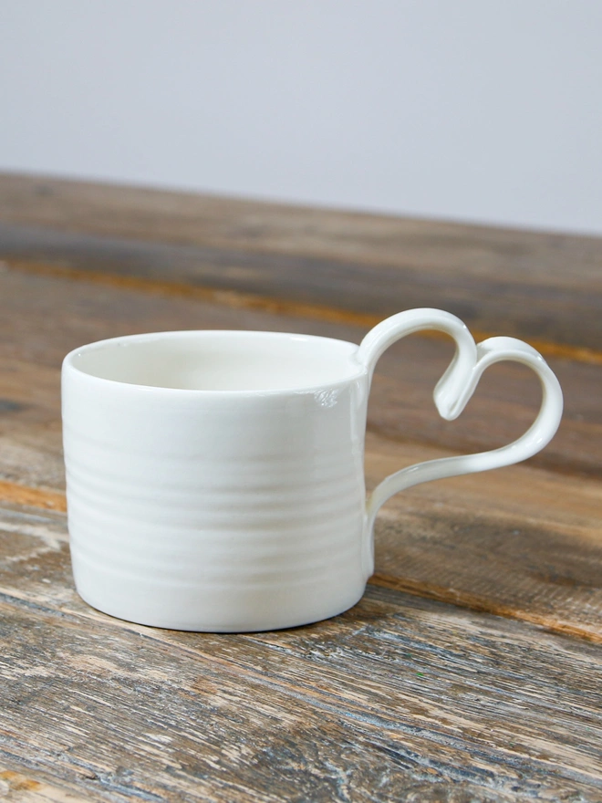 Heart shaped handle on mug in satin cream glaze sat upon a rustic table top