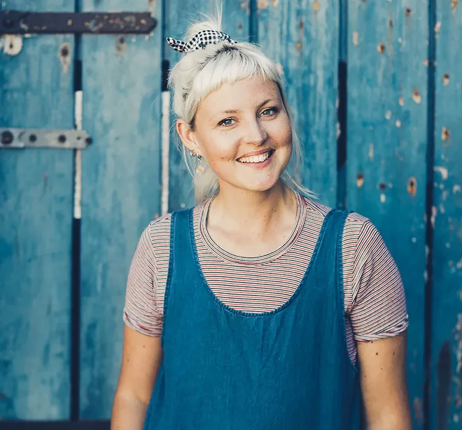 Kristin Hallenga, founder of CoppaFeel! smiling at the camera, stood infront of a blue wooden door.