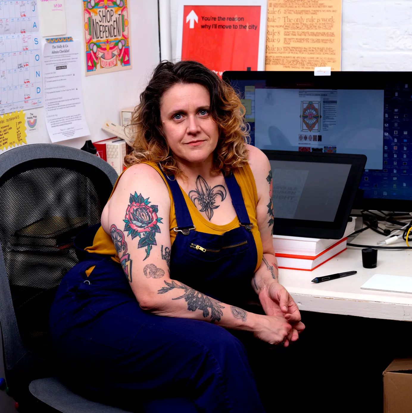 Picture show Rebecca sitting at her studio desk in Margate. There is a computer and drawing tablet just behind her and colourful pictures stuck to the white walls. On her desk is a pen, a candle, a phone and mousebad. Rebecca is wearing a yellow vest and royal blue dungerees. She has curly brown and blonde hair and many colourful tattoos.