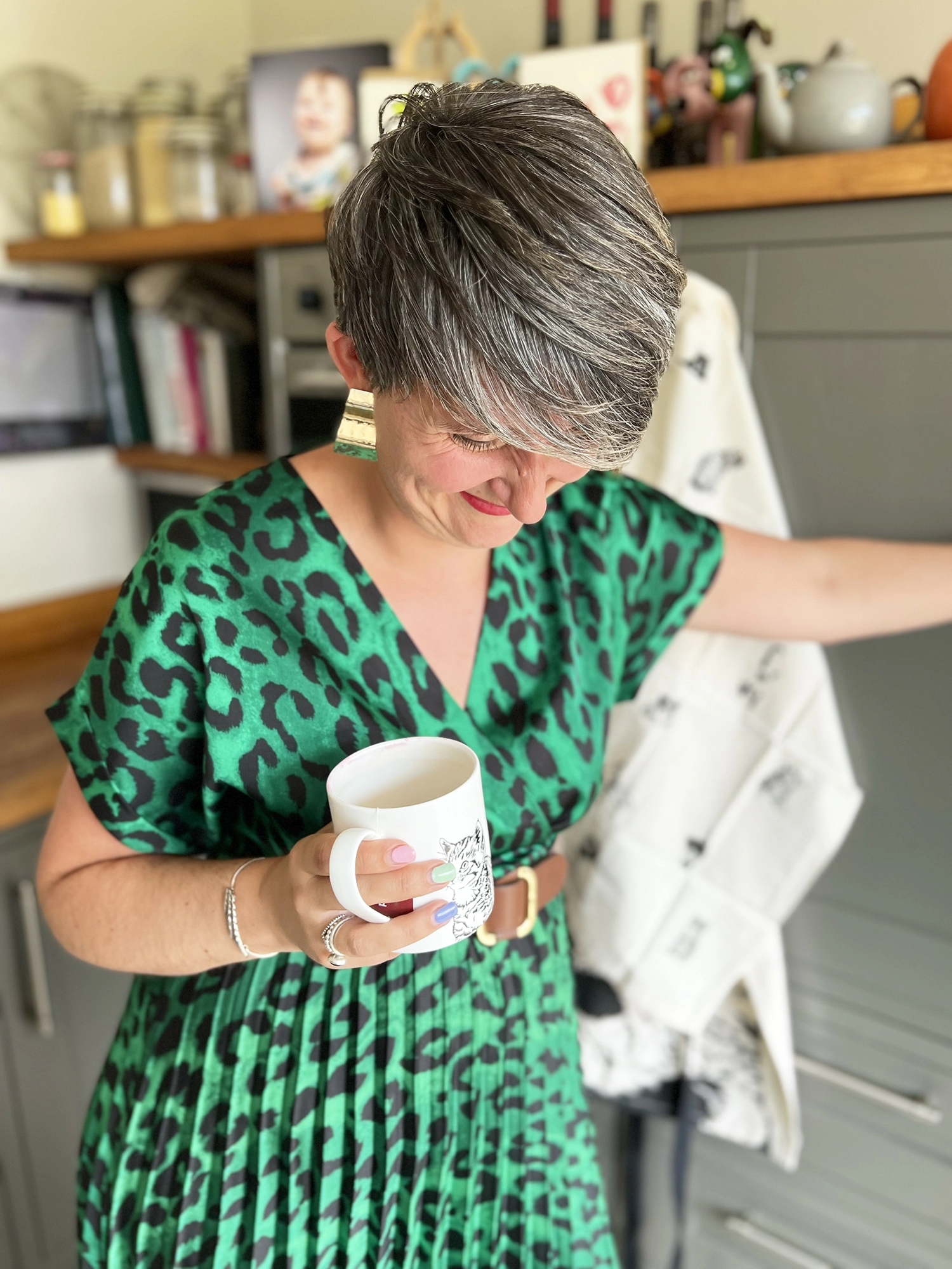 Photograph of Lizzie Parker the female founder of Penguin Ink standing in her kitchen, holding one of our Penguin Ink fine bone china mugs with tea towels in the background. She is laughing as she's not good at being serious in-front of a camera. 