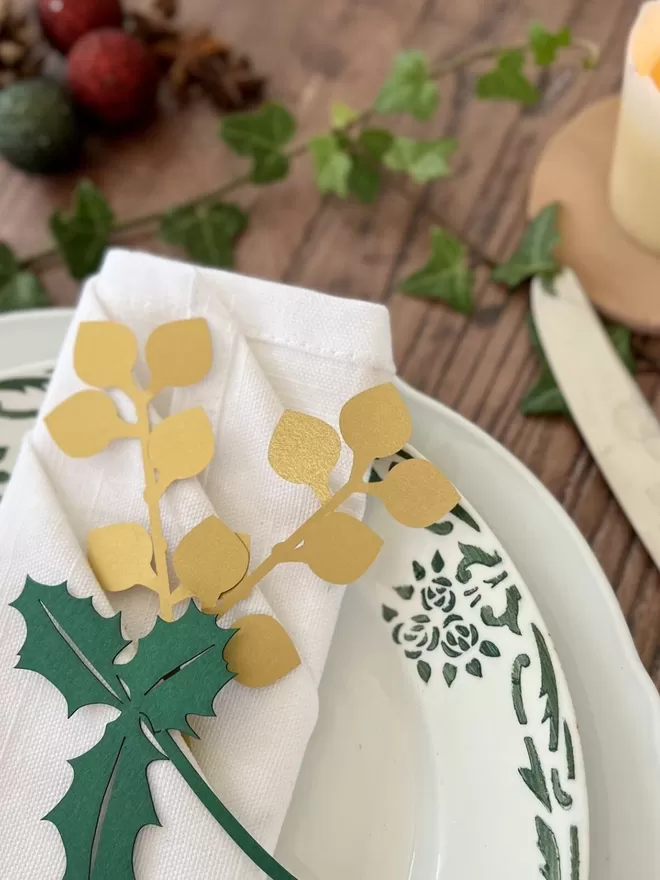 White folded napkind with gold and green winter foliage place setting decorations