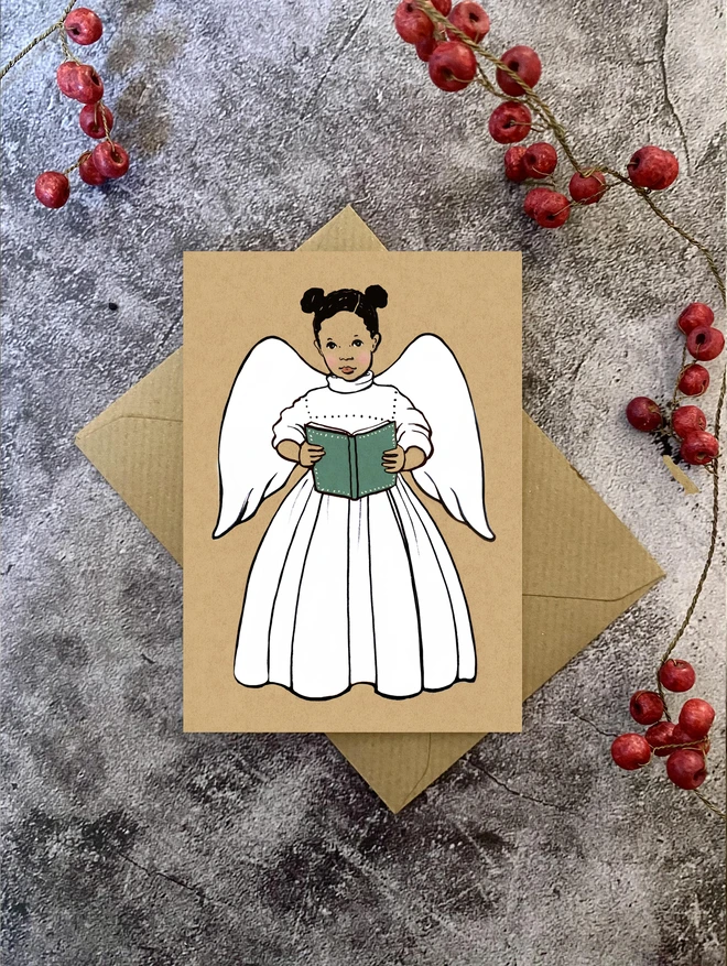 a greeting card featuring an afro Caribbean angel holding a blue song book card is laid on a Kraft envelope