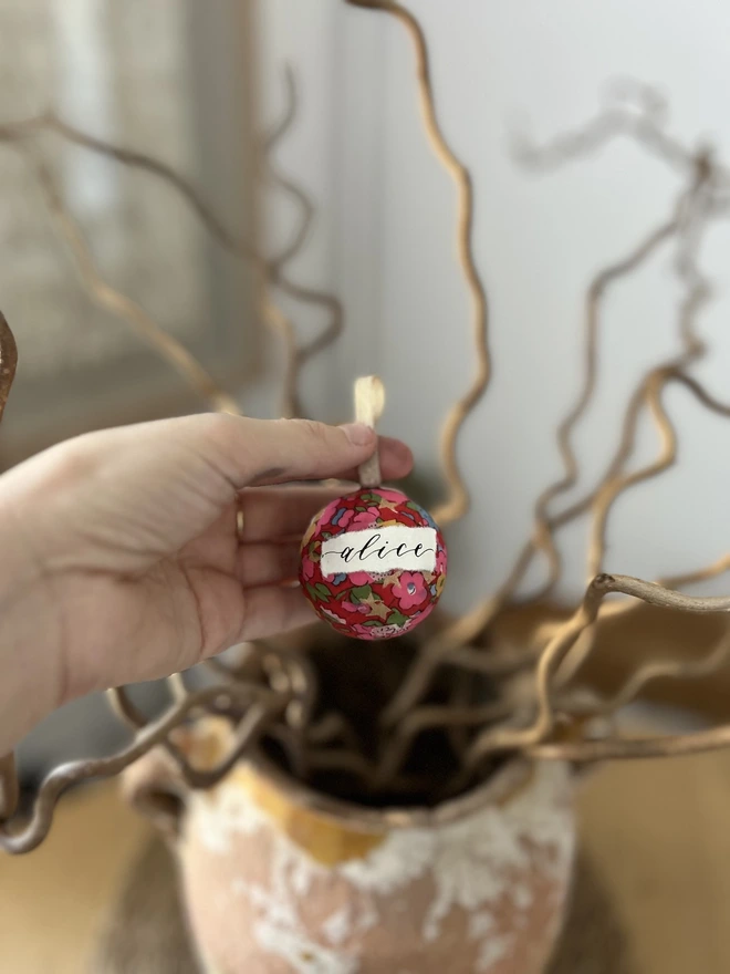 Mini Liberty fabric Christmas bauble with personalised Alice label held against a display of twigs  