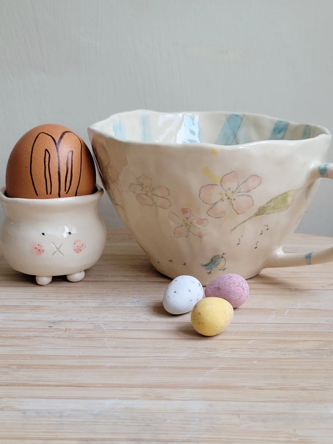 blossom and blue tit hand painted and hand made tea cup sitting next to a pottery easter bunny egg cup with an egg with bunny ears drawn on and mini chocolate eggs in the fore ground