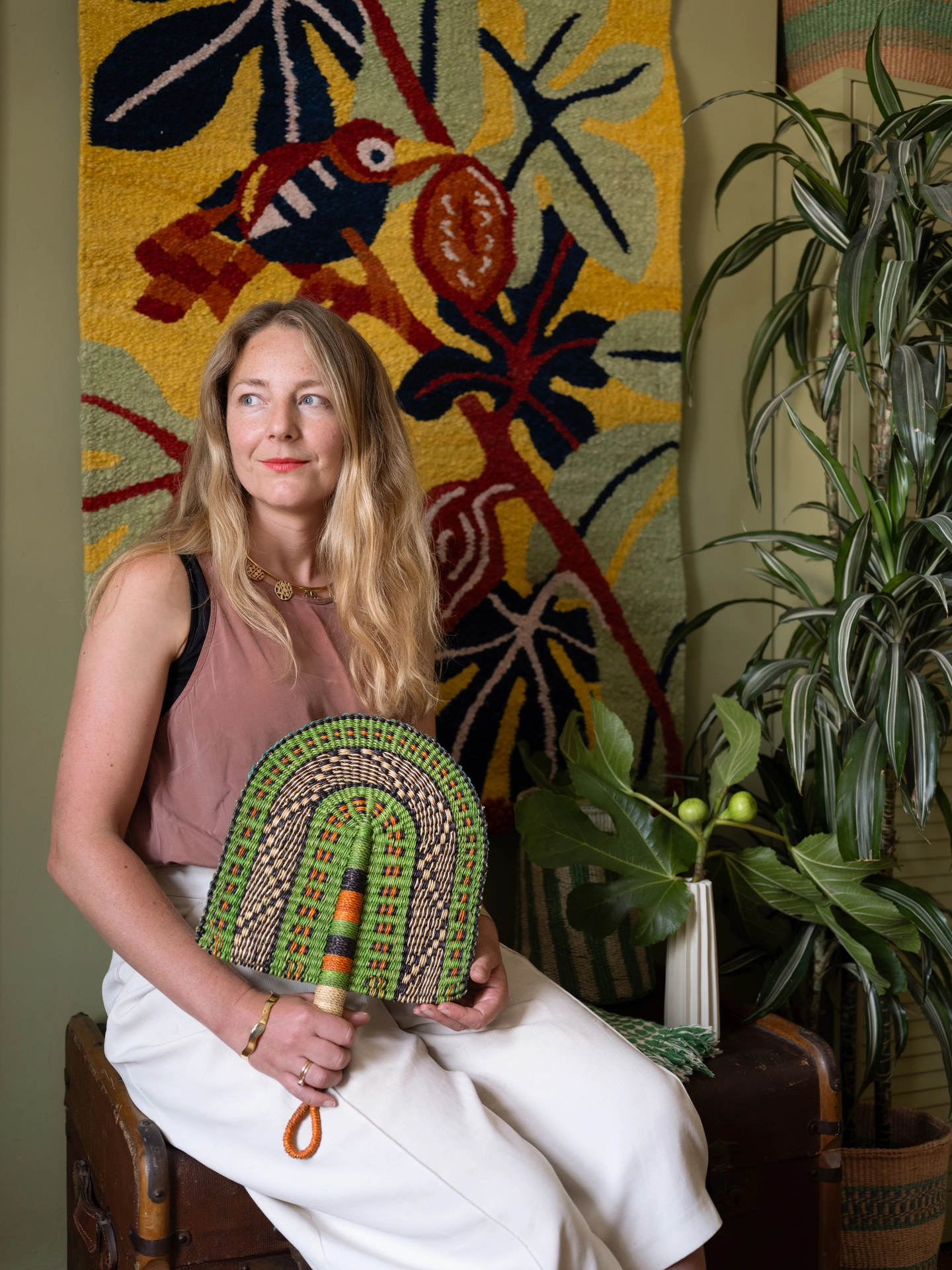 Amy, a white woman with blonde hair, sits in front of a woven wall hanging in green and yellow tones. The wall hanging features a bird motif and Amy is holding a green woven fan. 