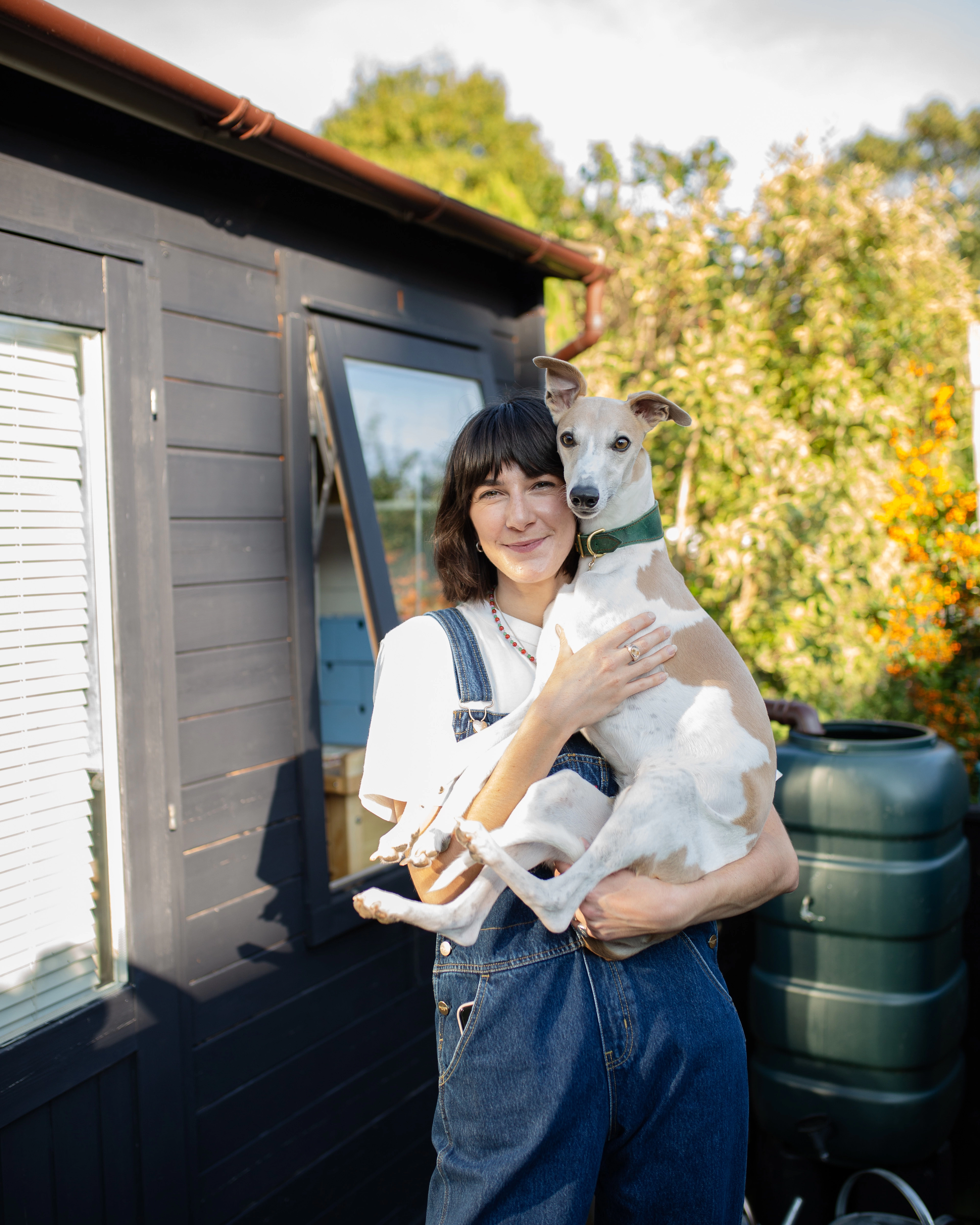 Brooke stands in front of her garden studio holding her whippet, Ralf.
