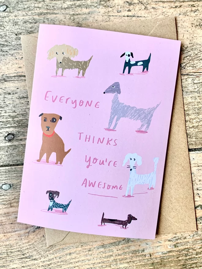 Card lies on wooden background with brown envelope. Pink card reads 'Everyone thinks you're awesome'