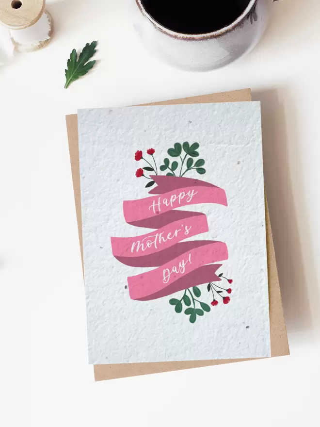 A card in the centre of the image placed on top of a brown envelope with a 'Happy Mothers Day' design on it. In the top of the image is part of a cup of coffee, a leaf and a roll of ribbon.