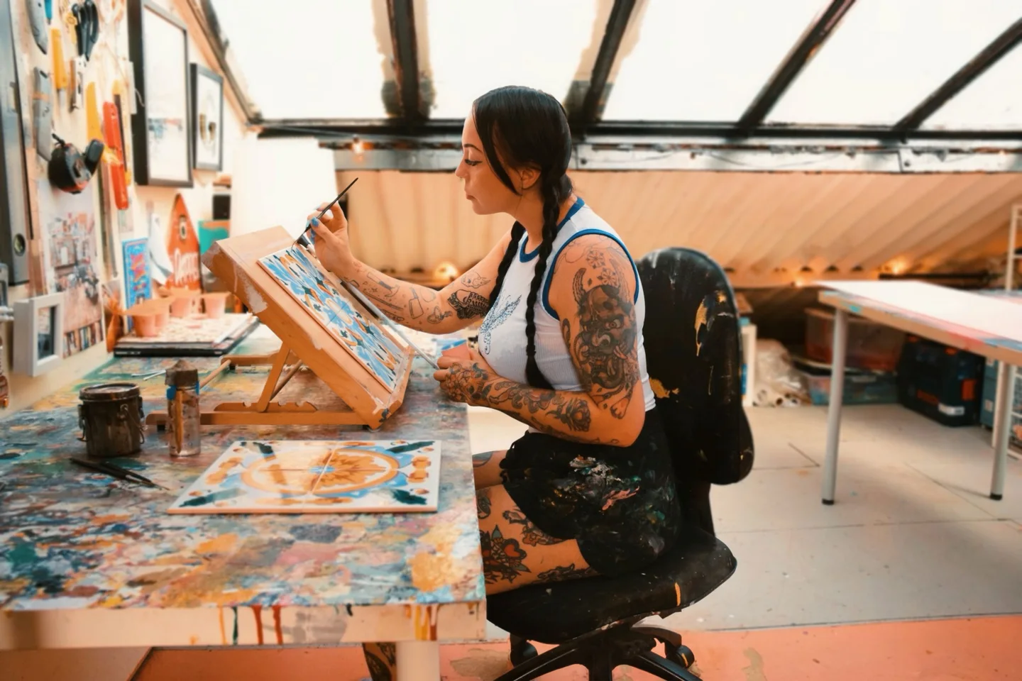 Tattooed woman in vest and shorts painting tiles with a mahl stick in hand. She is in a light and airy studio with paintings on the walls.