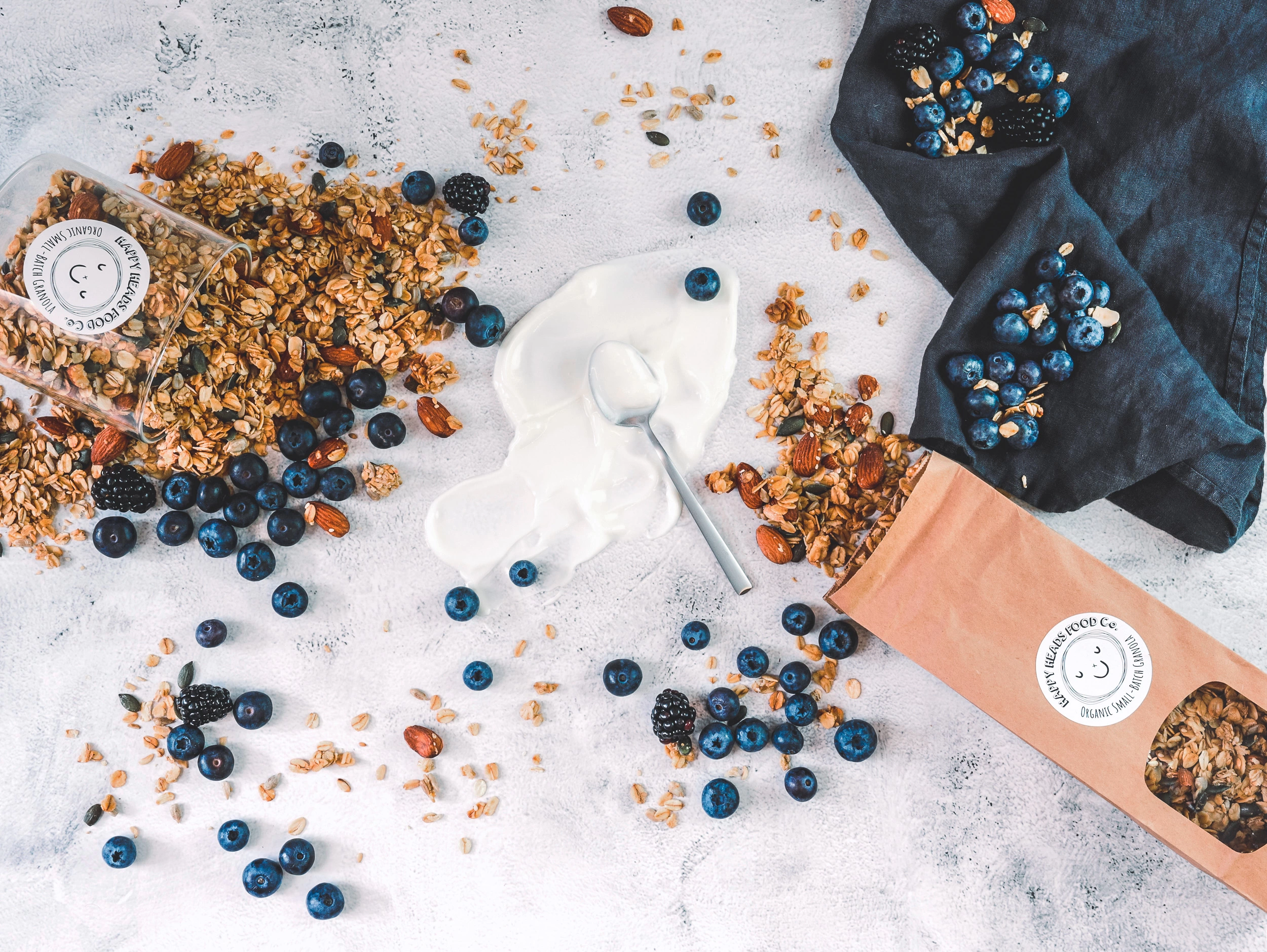 Happy Heads Food Co. 'Original' granola spilling from a glass jar and brown kraft paper bag on to a surface covered in yoghurt and blueberries.