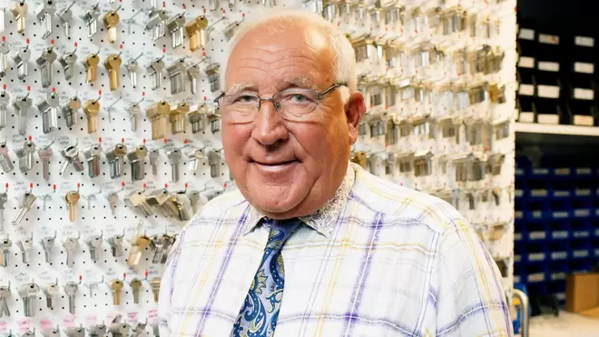 Sir John Timpson, chaiman & owner of Timpson, smiling at the camera stood infront of a wall of keys.