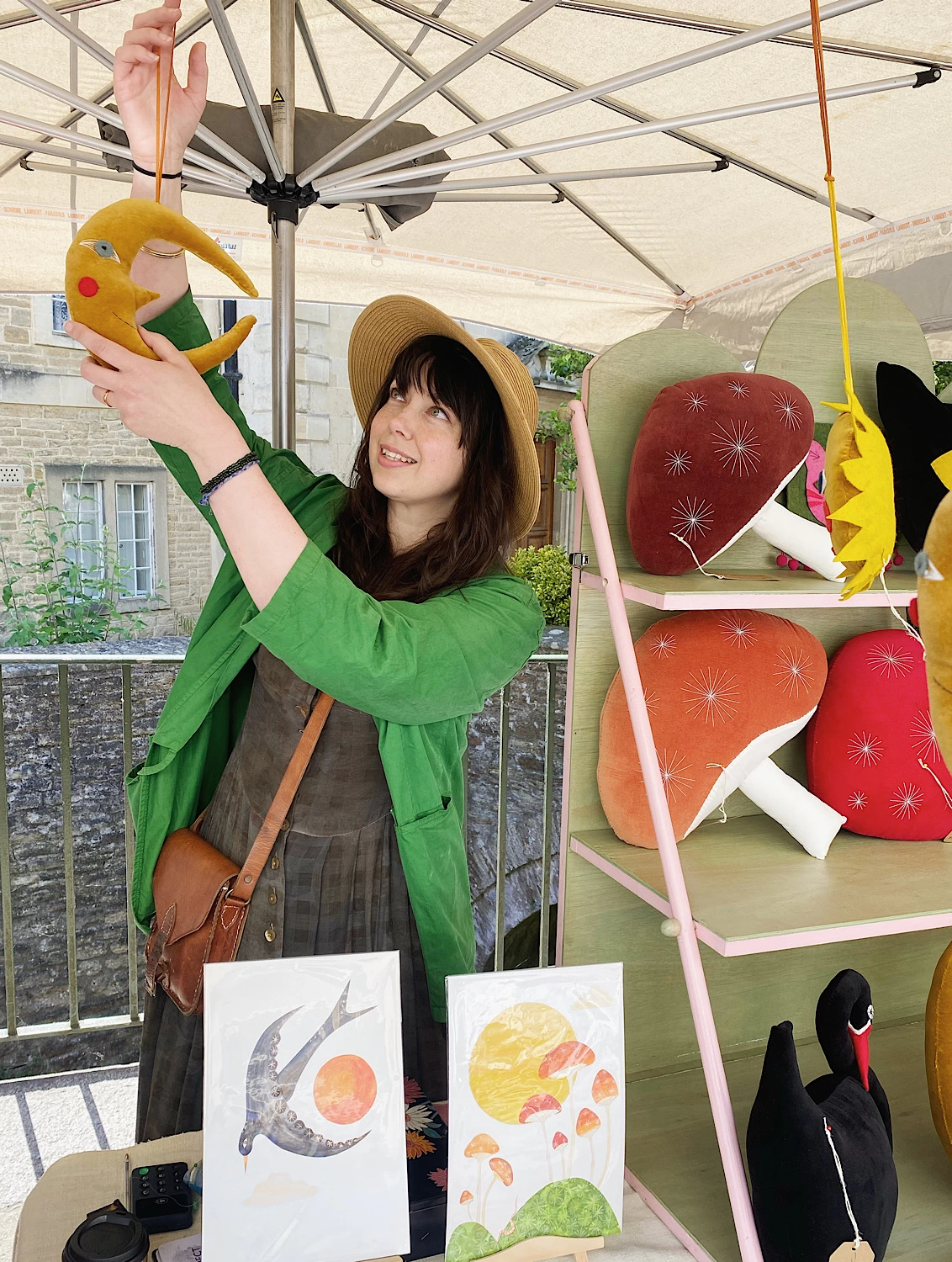 A dark haired, pale skinned woman in a hat and green jacket is standing behind a market stall full of brightly coloured cushions and artwork