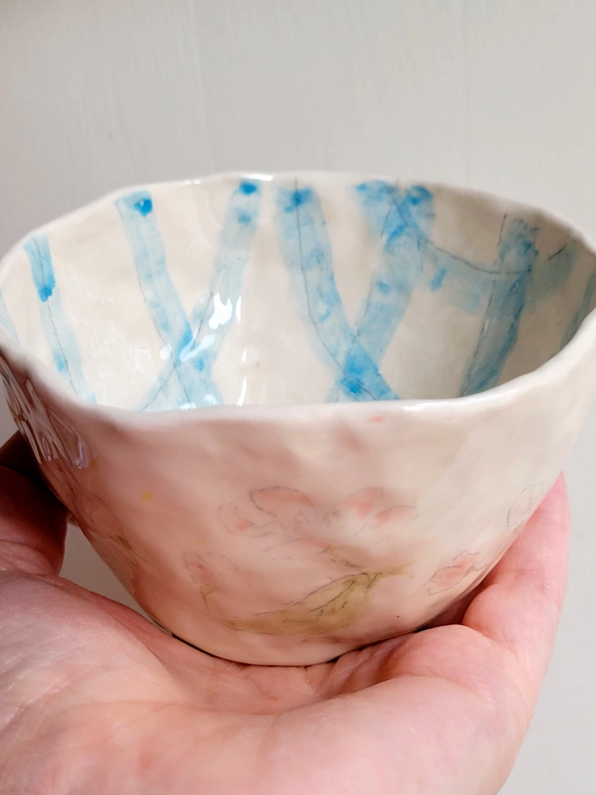 a ceramic tea cup with blue gingham check painted inside and pink flowers and green leaves on the outside in close up and held in a hand