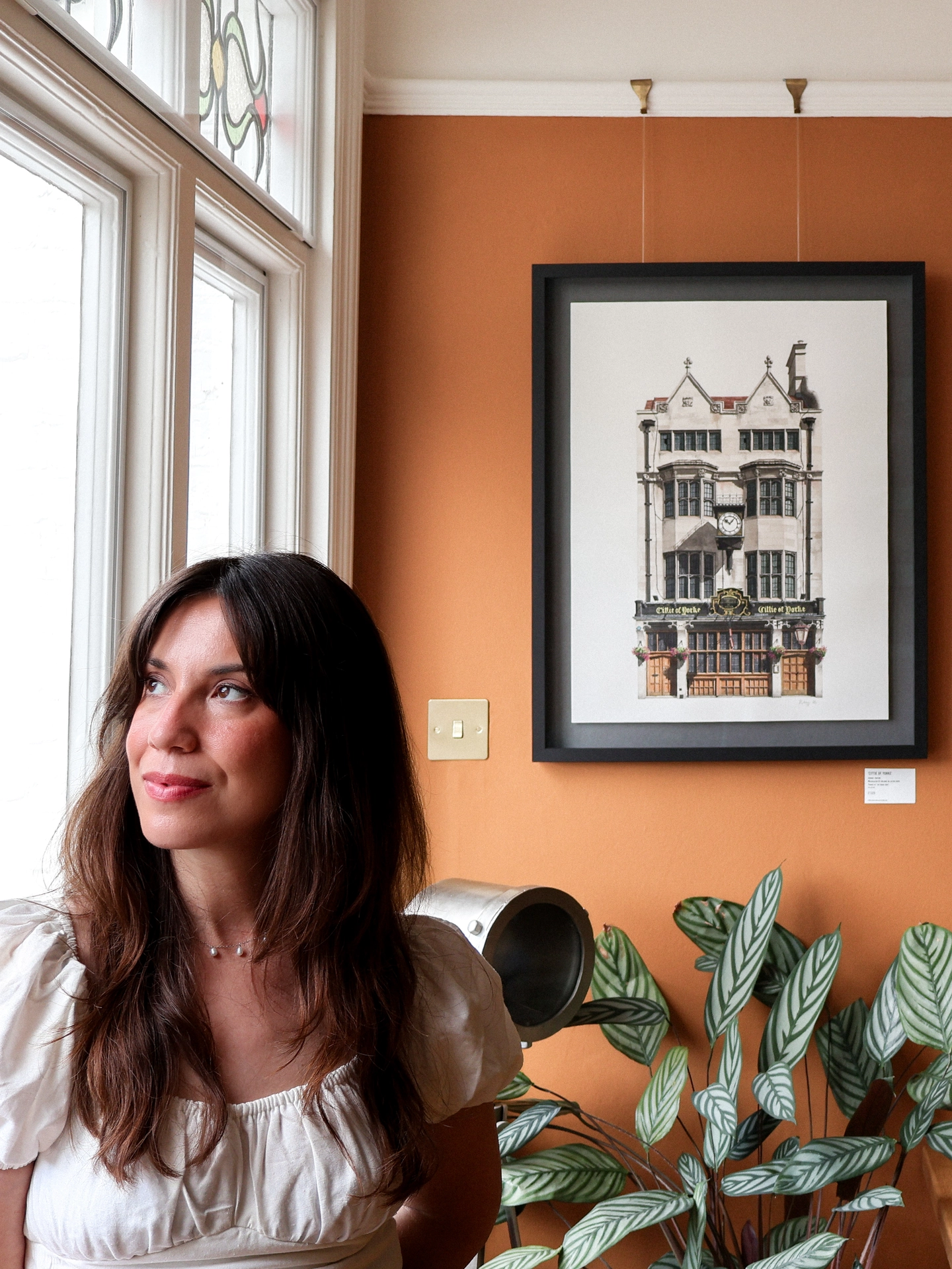 A woman looking out a window next to a framed watercolour print of the Cittie of Yorke pub hanging on an orange wall.