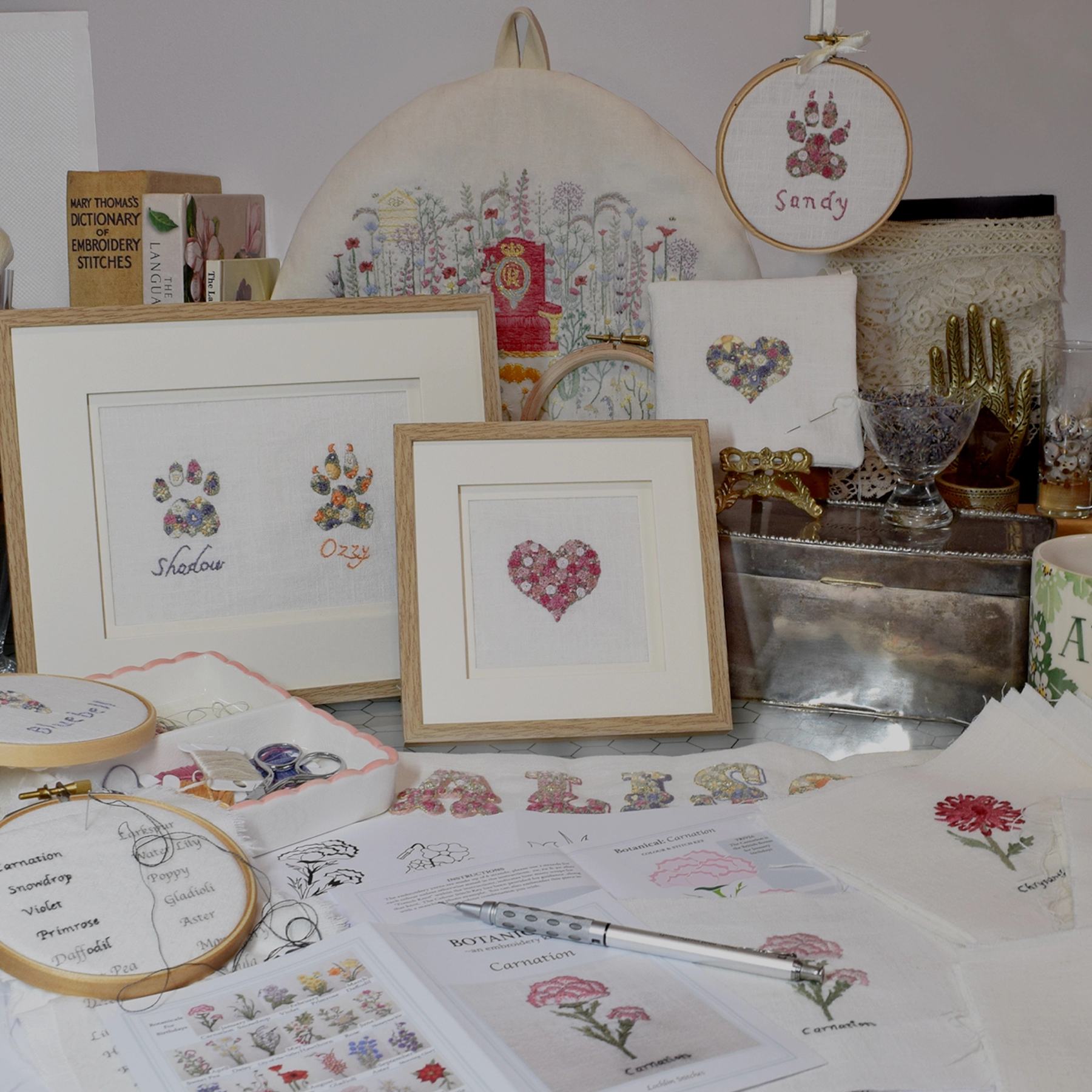 Lochlin Stitches, Designing Embroideries, In the Background are books on Flora and stitching, a King Charles III design, Dog Paw in a hoop, a heart lavender bag being made, and Desk ornaments.  In the Middle are Oak Framed embroidered Cat & Dog Paw, and a pink heart.  In the Foreground are Desgns in progress such as the Botanical Collection to be released later in 2024..