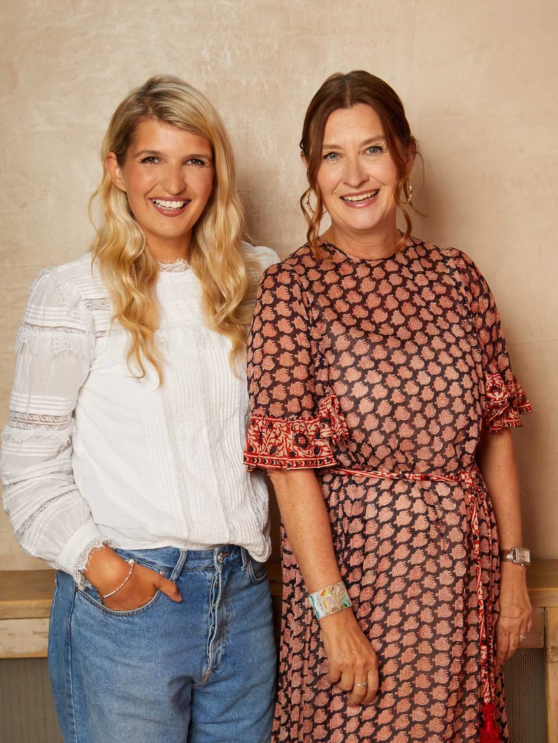 Designer, Sia with blonde hair, a white top and blue jeans stands with founder, Fiona of Their Nibs wearing a dark purple dress with pink small motifs and a tassel beltsmiling at the camera on a behind the scenes day at their AW'23 photoshoot.