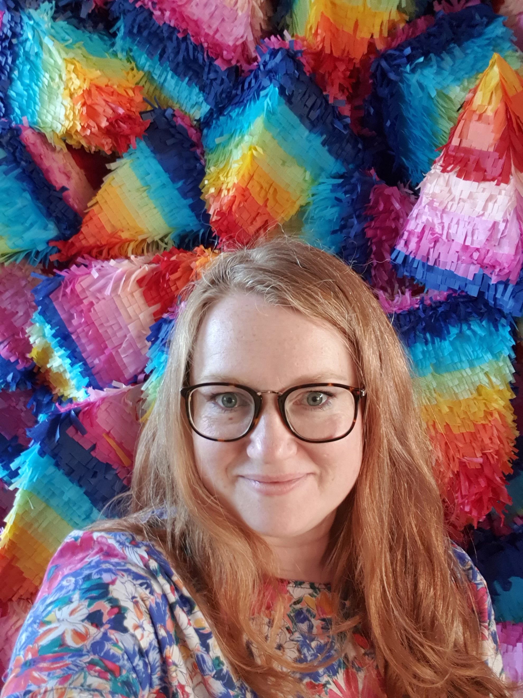 Portrait of Kate the face behind Pinyatay a white woman with glasses in front of a mountain of rainbow, diamond shaped pinatas.