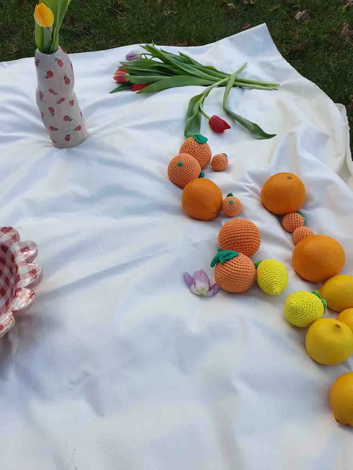 Crochet oranges and lemons on a white piece of fabric with handmade ceramics and tulips in the background.