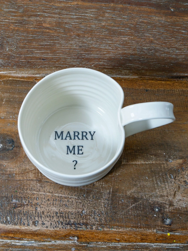 'Marry Me?' asked in black text on the inside of the hand thrown porcelain mug which sits atop a rustic wooden table top.