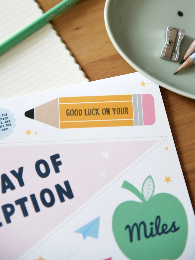  A "1st Day of Reception" card lies on a wooden desk surrounded by school supplies like scissors, pencils, a ruler, and a pencil case. The card includes a personalised name, encouraging messages, and playful designs.