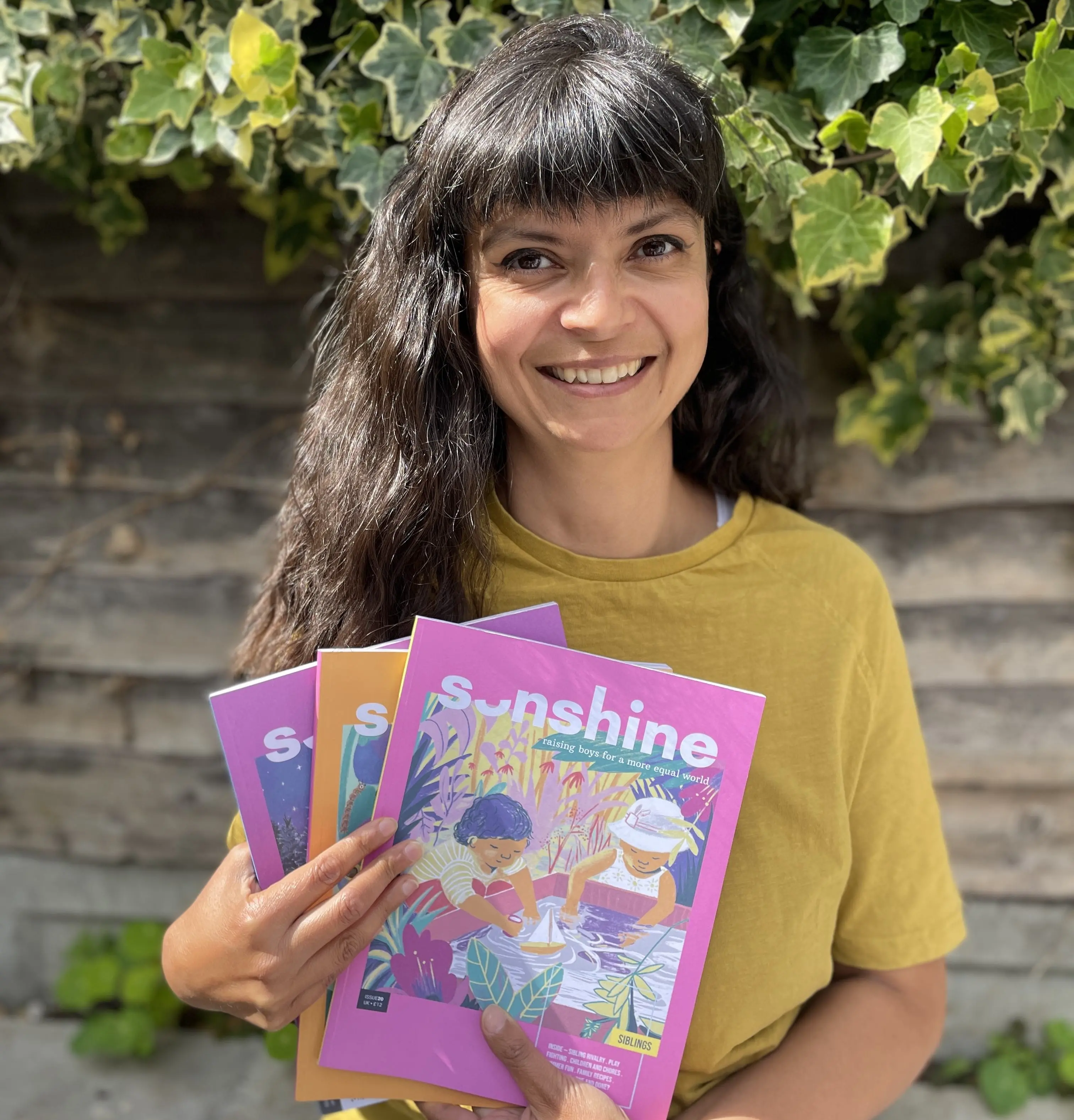 Kirstie wearing a yelllow shirt holding three issues of Sonshine Magazine against a garden background