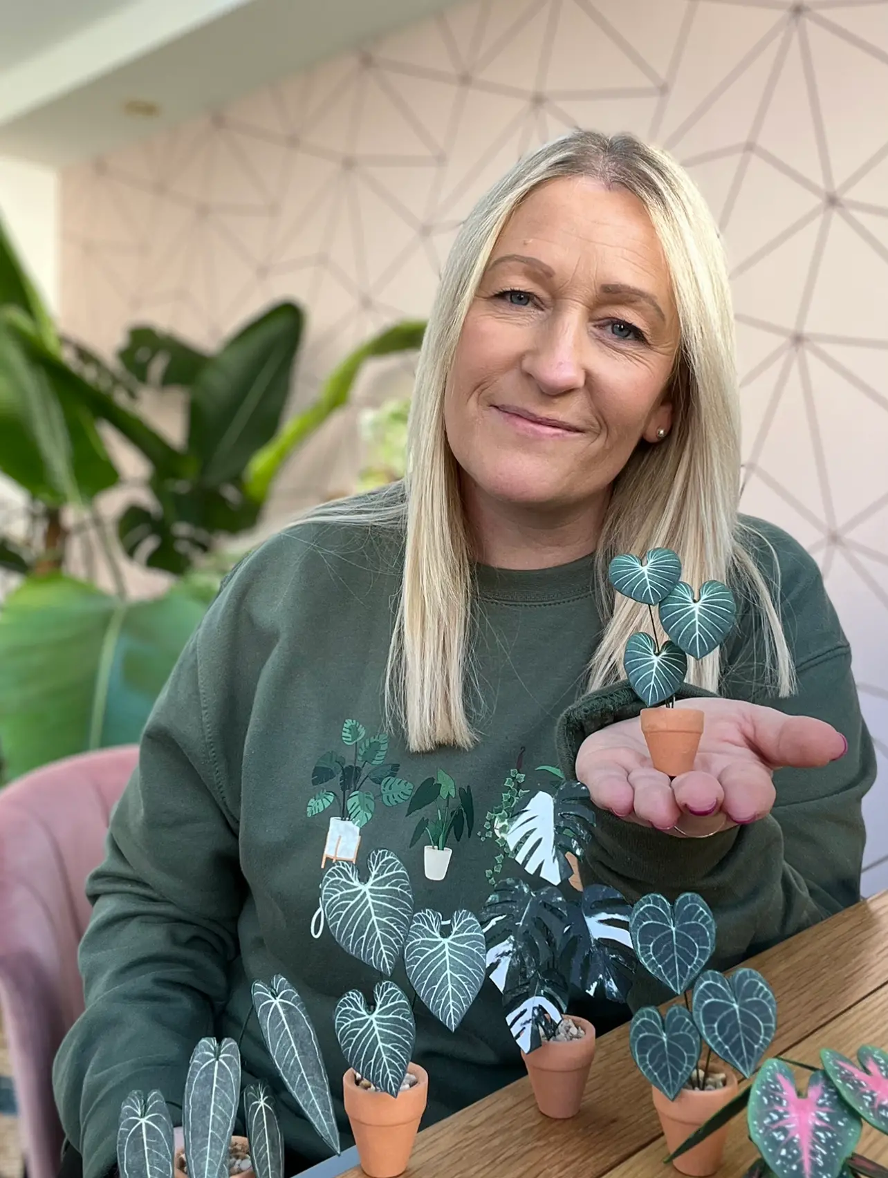 Nicole in her studio holding a miniature replica Philodendron Gloriosum paper plant ornament in a terracotta pot on the palm of her hand with other paper plants in the foreground