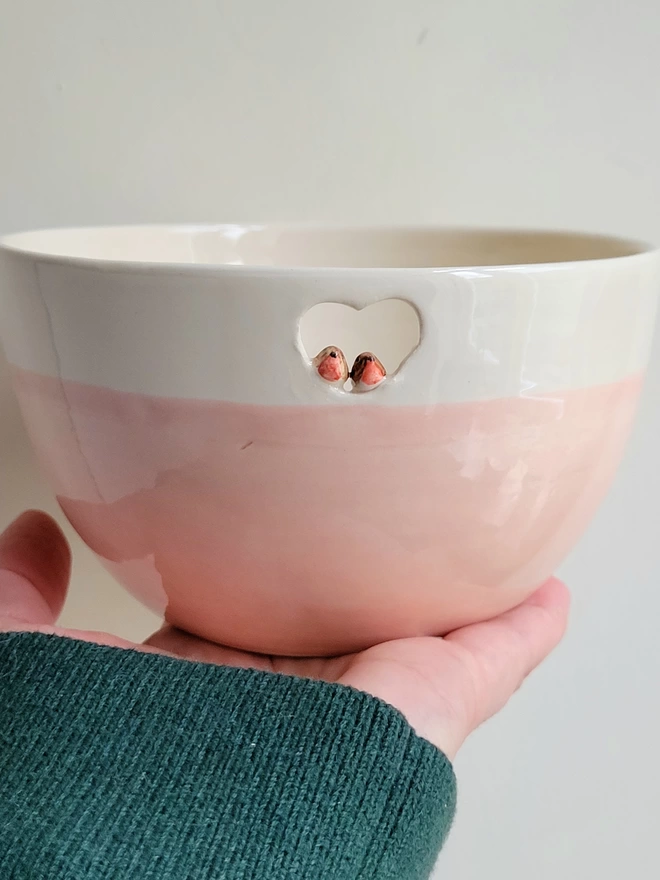 a pink pottery bowl being held in a hand. there are two tiny ceramic robin birds perched inside a cutout heart near the rim of the bowl