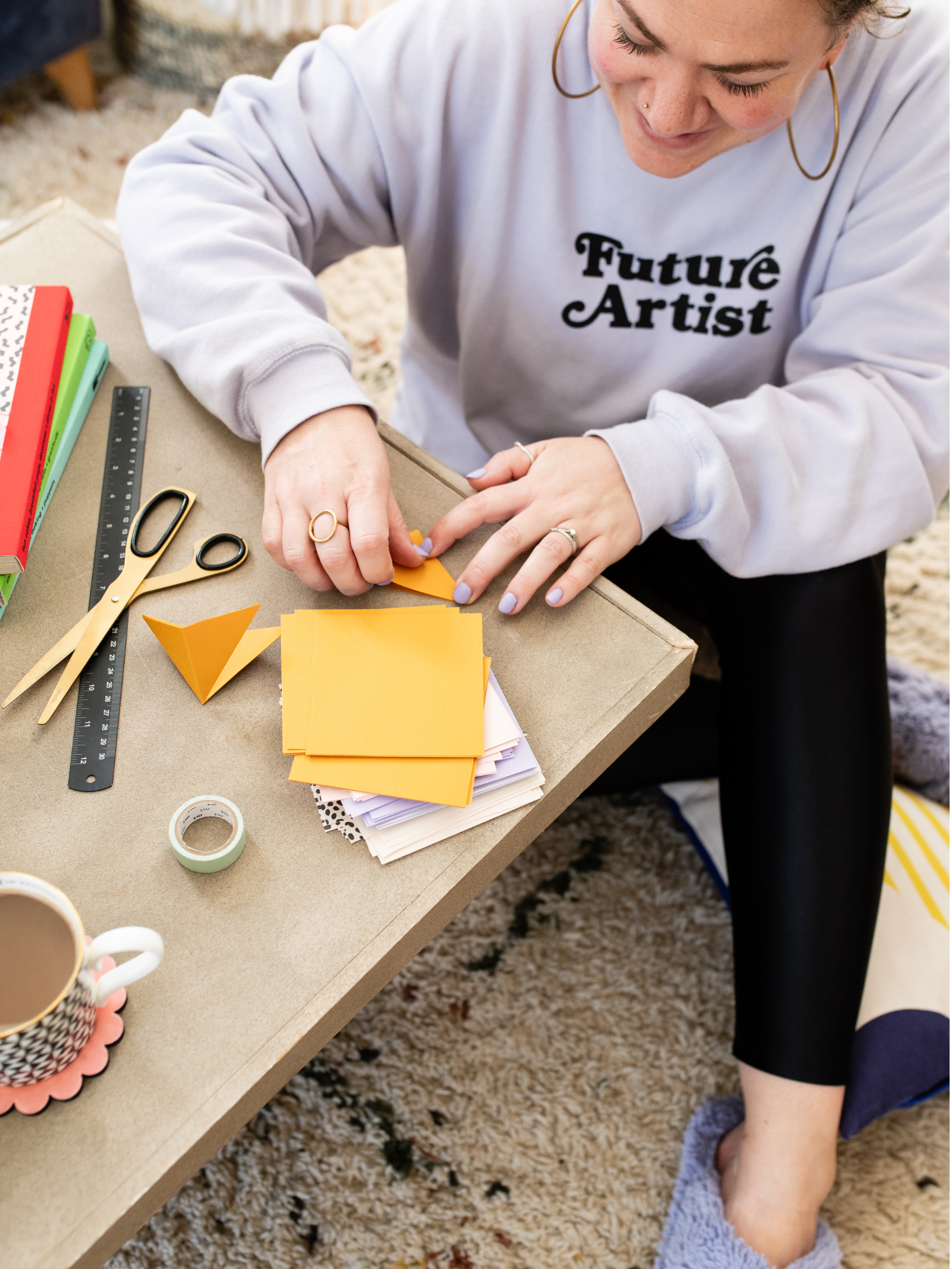 Woman is folding paper squares on a low rise table, with scissors and tools to hand. She's smiling as she folds.