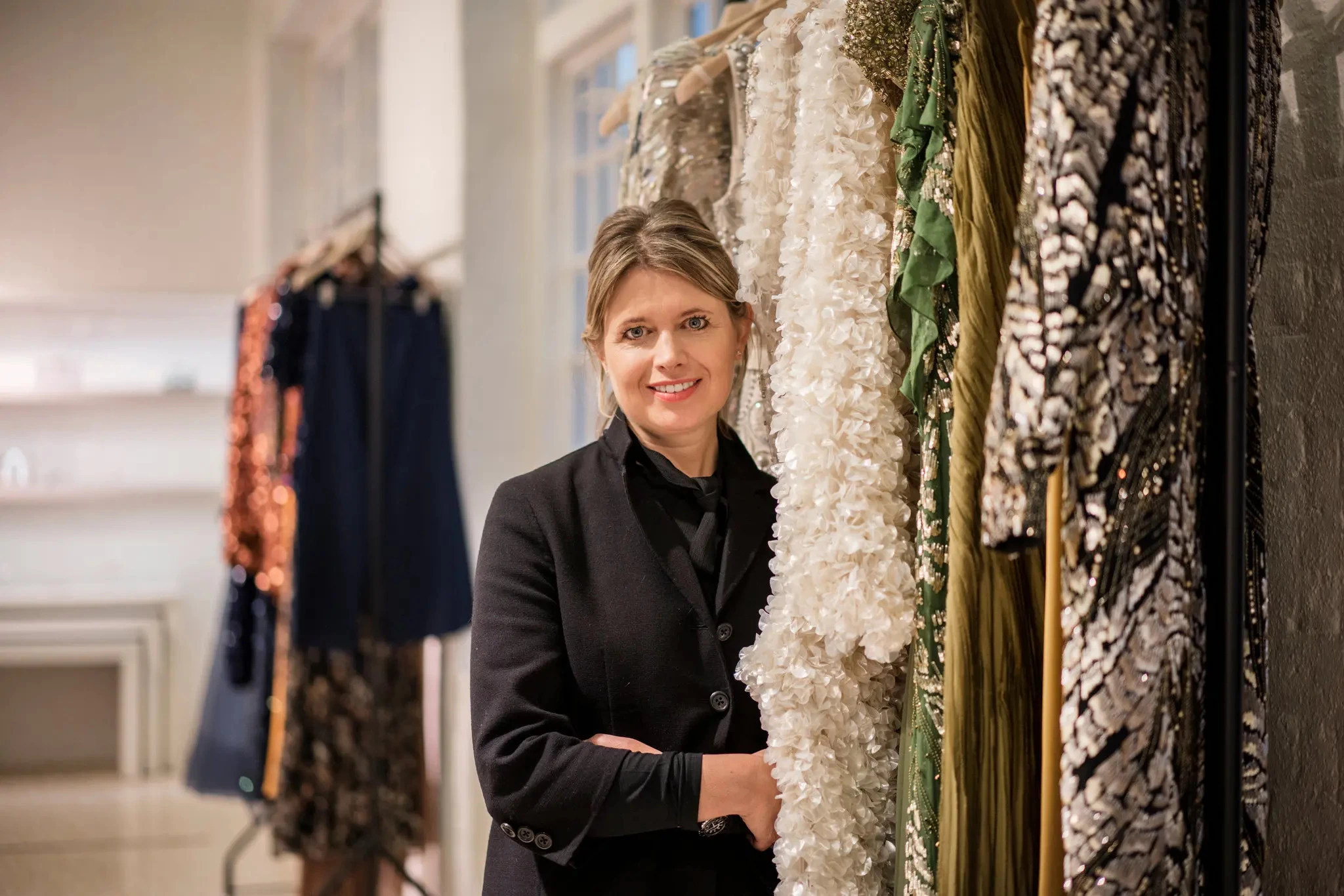 Jenny Packham, designer and founder of Jenny Packham, smiling at the camera, stood next to a rack of Janny Packham dresses.