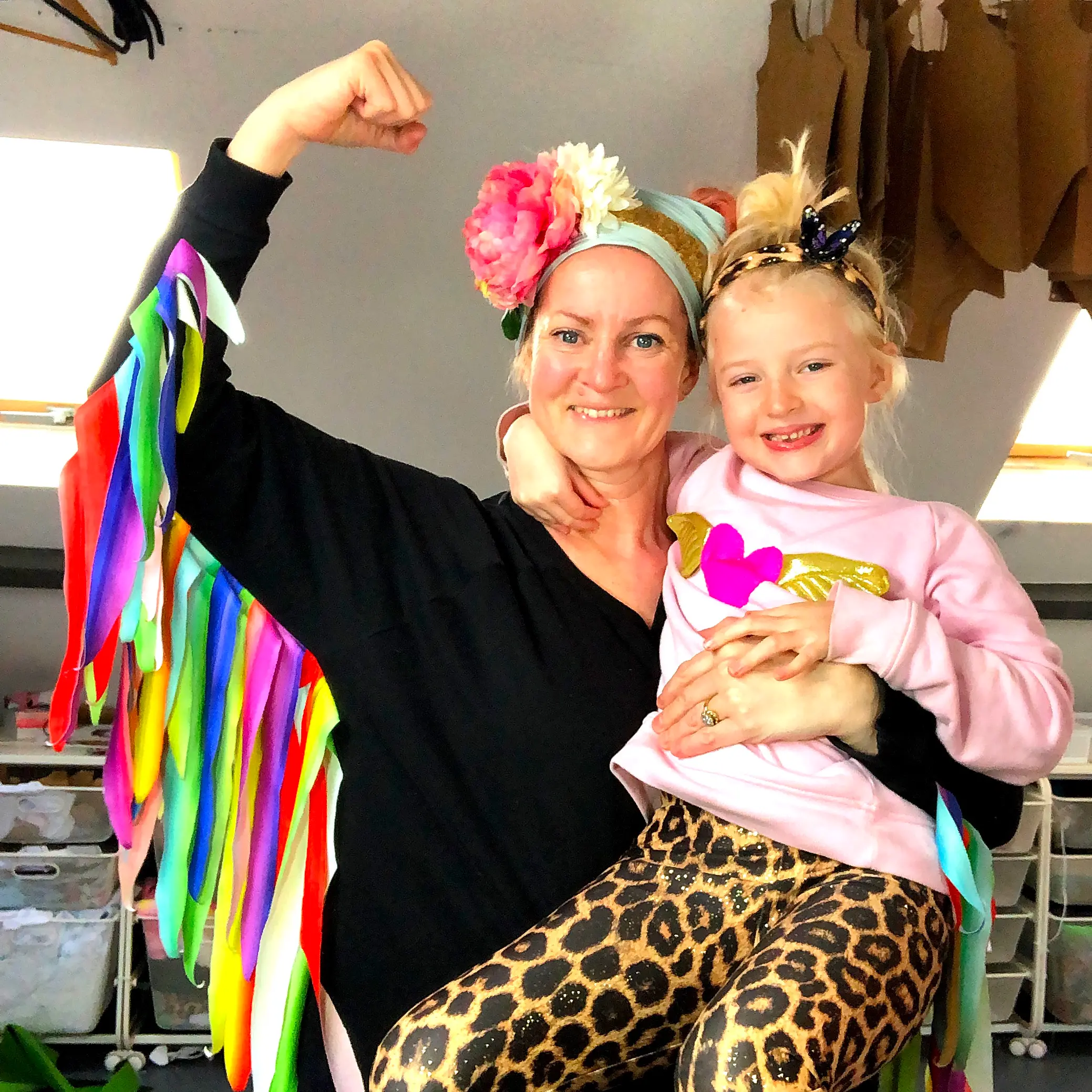 Founder of childrens brand 'Flingo and Swollop' standing in her studio with her daughter. both wearing Flingo and swollop clothes. A black sweatshirt with rainbow wings. Her daughter is wearing a pair of sparkly leopard print leggings. they are both smiling.
