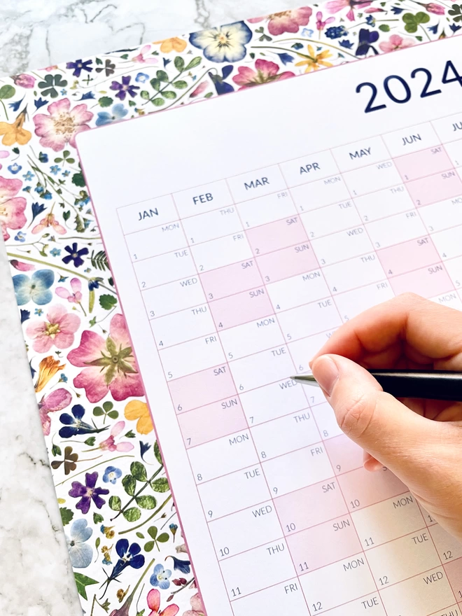 A Hand Holding a Pen Ready to Write on a Yearly Wall Planner with a Floral Border