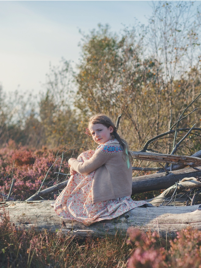 A girl in a beige cardigan with a fair isle yoke over a floral dress sits on a log surrounded by heather